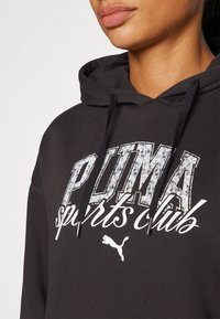 Felpa nera con cappuccio caratterizzata da un grande logo "PUMA sports club" bianco, consumato, e da un piccolo emblema Puma. Il materiale sembra morbido con una finitura testurizzata.