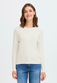 Cardigan crème clair avec un motif de tricot texturé, manches longues et fermeture à boutons. Associé à un pantalon en denim bleu.