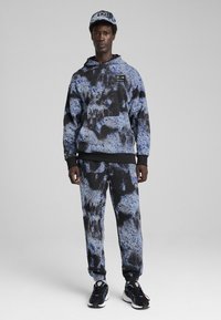 Sudadera con capucha y pantalones de chándal en un patrón de tie-dye azul y negro, con acentos negros y un parche con logo, combinados con zapatillas negras.