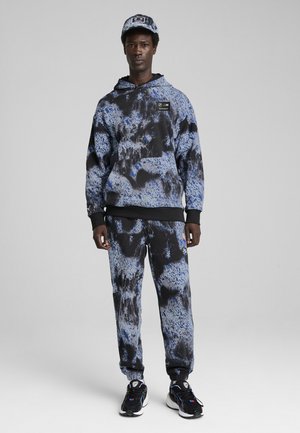 Hættetrøje og sweatpants i blå og sort tie-dye mønster, med sorte detaljer og et logo-patch, sammen med sorte sneakers.