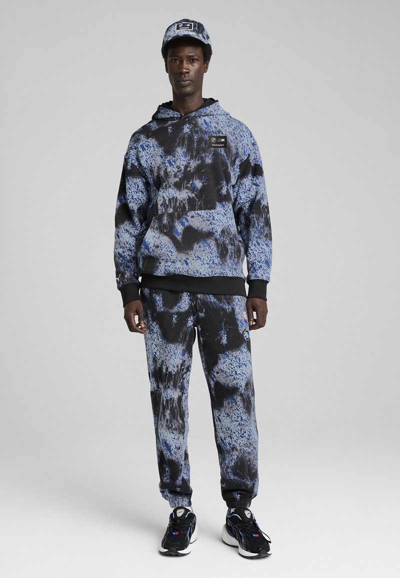 Sudadera con capucha y pantalones de chándal en un patrón de tie-dye azul y negro, con acentos negros y un parche con logo, combinados con zapatillas negras.