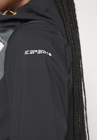 Veste noire avec un panneau gris texturé sur l'épaule. Présente un cordon orange et le logo blanc "ICEPEAK" sur la manche.