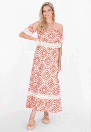 Giovane donna sorridente, indossa un abito lungo scollato con motivo geometrico rosso e arancione, rifinito con pizzo, e sandali beige con zeppa.