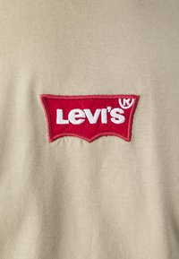 Beige stof med Levis logo i rød broderi, der har en syet patch kant. Glat tekstur med synligt strikkemønster.