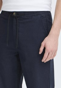 Navy blue pants with a drawstring waist, featuring a button closure and side pockets. Material appears textured with a relaxed fit.