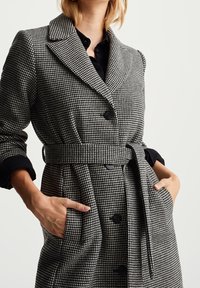 Cappotto a fantasia pied de poule grigio e nero con vita con cintura, grandi risvolti e bottoni neri. Caratterizzato da tasche laterali e una vestibilità sartoriale.
