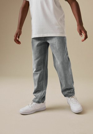 Person trägt lockere graue Jeans mit elastischen Bündchen und weißen Sneakern, steht vor einem einfarbigen beigen Hintergrund.