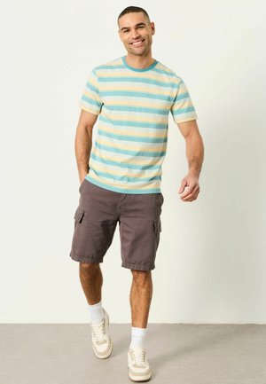 Smiling man walking forward wearing a pastel striped short-sleeve shirt, brown cargo shorts, white socks, and beige sneakers against a white background.