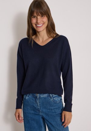 Smilende kvinde med brunt hår iført en mørkeblå langærmet sweater og blå jeans står foran en ensfarvet lys baggrund.