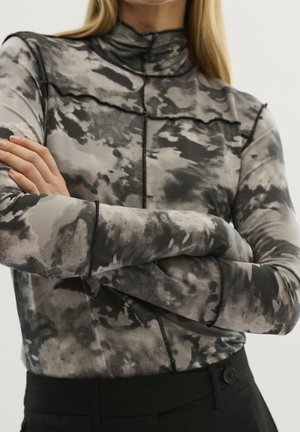 Personne portant un haut à manches longues et col montant avec un motif abstrait noir et gris et un pantalon noir, les bras croisés sur la poitrine.