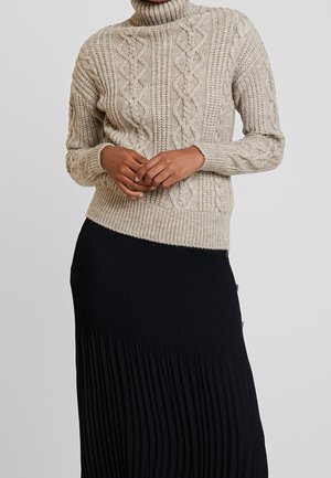 Personne portant un pull à col roulé en maille torsadée beige et une jupe plissée noire, debout devant un fond gris uni.