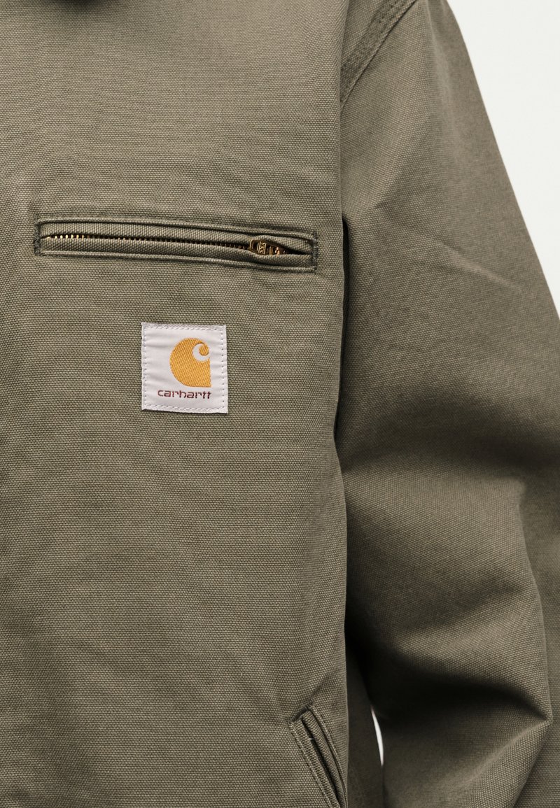 Primo piano di una giacca Carhartt verde oliva con tasca sul petto con cerniera e patch con logo del marchio visibile sul tessuto.