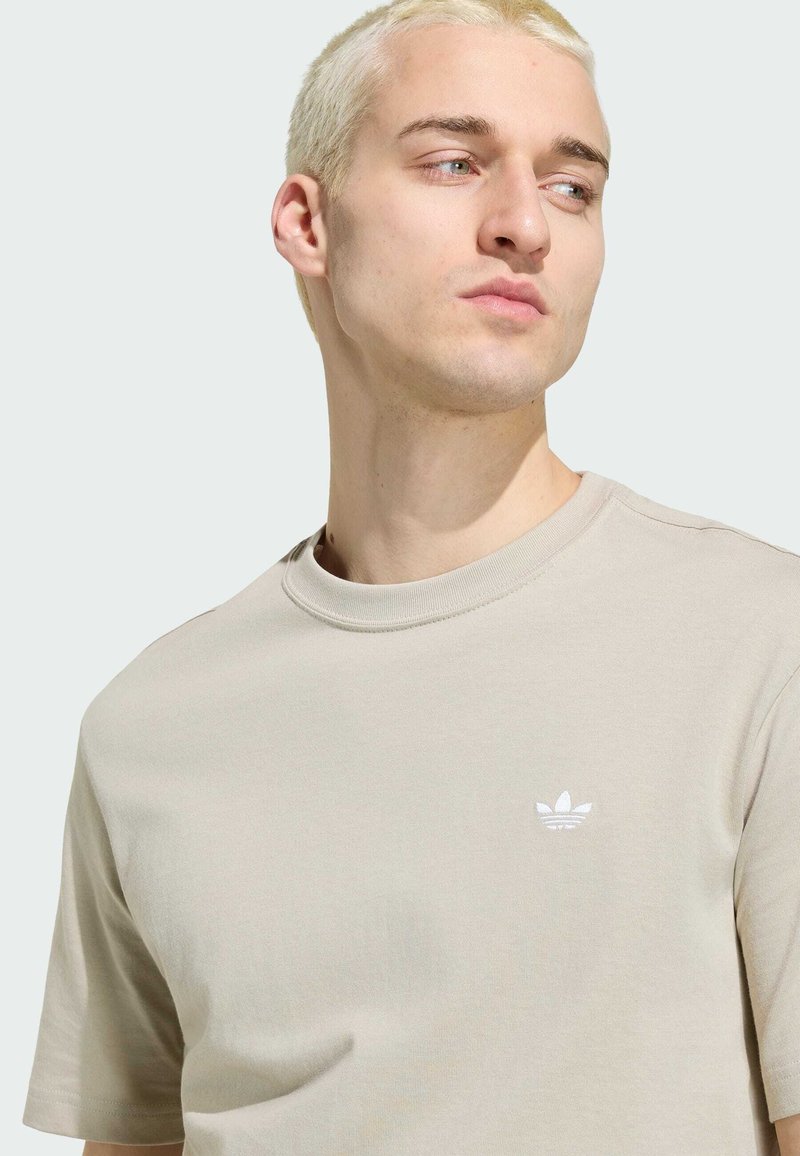 Blonde man kijkt naar rechts, draagt een beige T-shirt met ronde hals en een klein wit Adidas-logo op de borst tegen een lichte achtergrond.