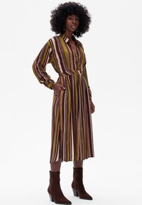 Pasiasta sukienka midi w kolorach brązowym, zielonym i beżowym. Posiada długie rękawy, zapinanie na guziki z przodu oraz plisowany dół. Zestawiona z brązowymi botkami za kostkę.
