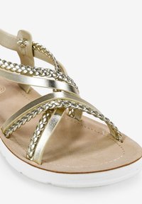 Sandal de metal dorado con tiras entrelazadas, plantilla acolchada en tono beige y suela blanca. Las tiras cruzadas aseguran un ajuste seguro.