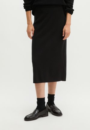 Personne portant une jupe midi noire, des chaussettes noires et des mocassins noirs brillants, debout sur un fond clair.