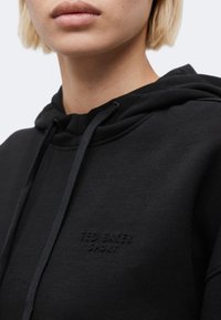 Κοντινό ενός ατόμου που φοράει μαύρη φούτερ με κουκούλα, με κεντημένο το λογότυπο "Ted Baker Sport" στο στήθος, διαθέτει κορδόνια και κουκούλα.