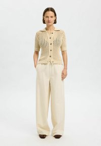 Cárdigan de punto beige con mangas cortas, cierre de botones y un patrón texturizado. Combinado con pantalones sueltos de color claro.