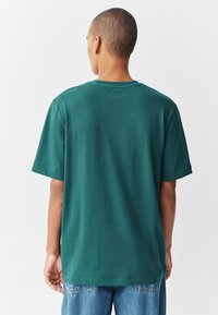 Žalias smaragdinis medvilninis marškinėlis trumpomis rankovėmis ir apvalia apykakle. Turi laisvą siluetą ir švelnią tekstūrą; be matomų logotipų ar grafikos.