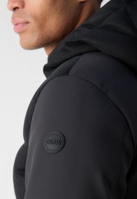 Black hoodie with textured hood, matte fabric, and a circular logo patch on the left shoulder. Features a smooth finish and athletic design.