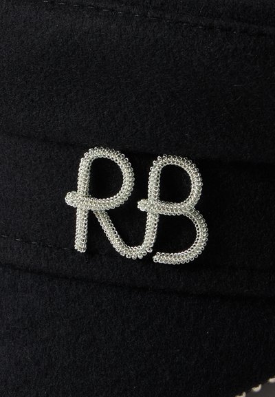 Ornement en perles argentées en forme de lettres "RB" sur un fond en tissu noir texturé.