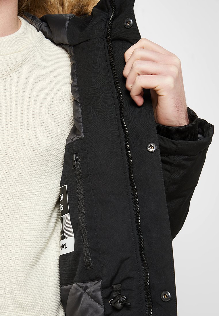 Una persona con un suéter blanco texturizado sostiene abierta una chaqueta de invierno negra, revelando un bolsillo interior con cremallera y una etiqueta.