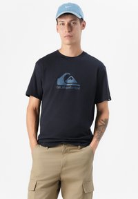 Jaunas vyras su trumpais plaukais dėvi tamsiai mėlyną Quiksilver marškinėlį ir smėlio spalvos krovinių šortus, stovintis prieš baltą foną su rankomis kišenėse.