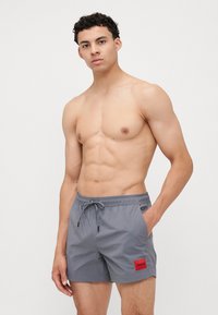 Shorts de bain gris avec une taille élastique et un cordon de serrage. Comprend des poches latérales et un patch logo rouge sur la jambe gauche. Texture lisse.