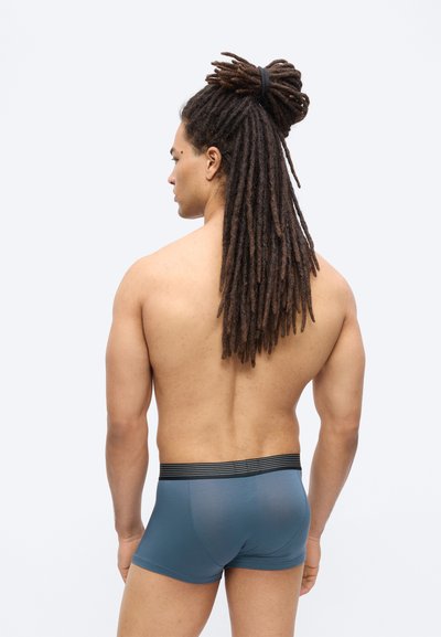 Άνδρας με μακριές κοτσίδες δεμένες πίσω, χωρίς μπλούζα, φοράει μπλε μπόξερ, στέκεται και κοιτάζει προς τα πίσω με φόντο ένα απλό λευκό φόντο.