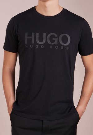 Osoba w czarnej koszulce HUGO BOSS z dużym logo "HUGO" i rękami w kieszeniach, pokazana od szyi do pasa.