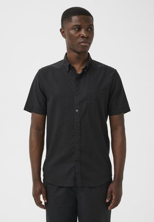 Homme adulte faisant face à l'avant, regardant vers la droite, portant une chemise noire à manches courtes avec une poche poitrine, debout devant un fond clair uni.