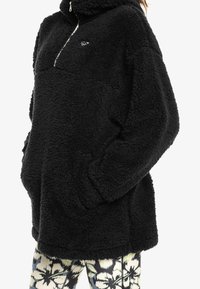 Zwarte fleece pullover met een hoge kraag en een halve rits aan de voorkant. Voorzien van zakken aan de voorkant en een gestructureerd, pluizig oppervlak.