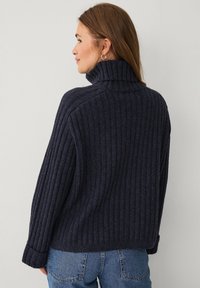 Navy ribbed knit sweater with a high collar and relaxed fit. Features long sleeves and folded cuffs, paired with blue denim jeans.