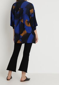 Blå og sort mønstret kimono-lignende top med brede ærmer og løs pasform, parret med sorte vide bukser og slip-on sko.