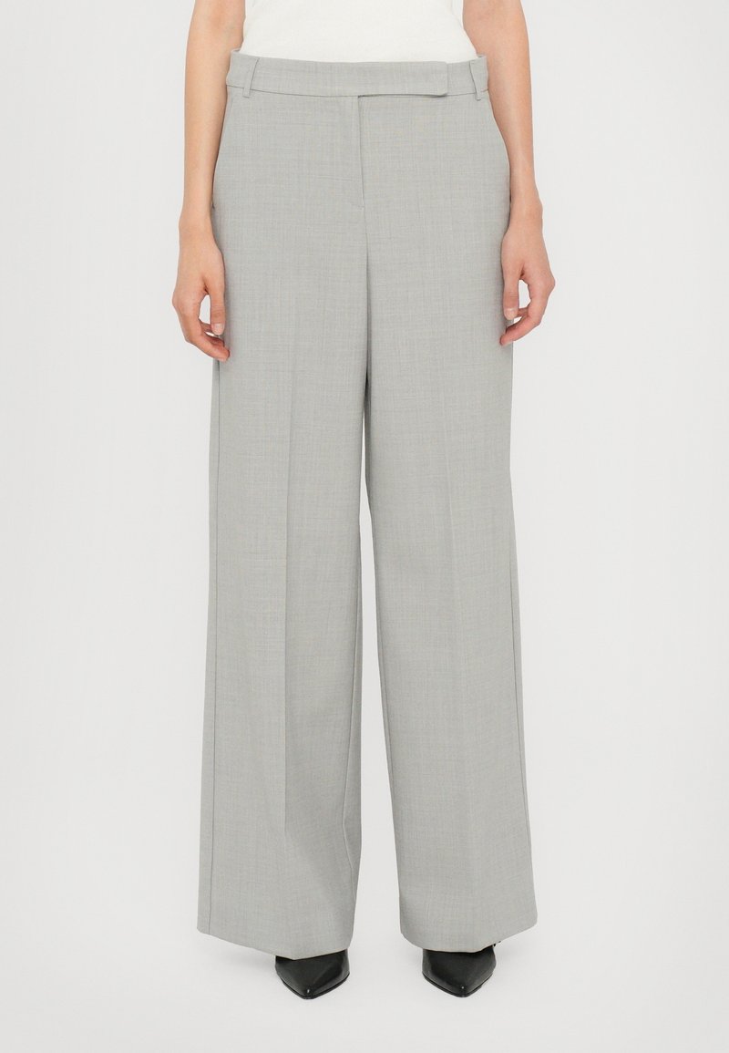 Femme portant un pantalon large gris clair avec des plis sur le devant et une fermeture de taille dissimulée, associé à des chaussures noires à bout pointu.