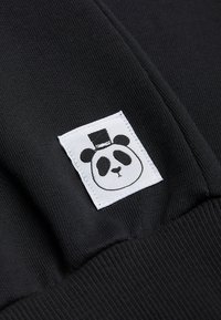 Tela negra con un parche rectangular blanco que muestra la cara de un panda usando un sombrero de copa negro, cosido cerca de un puño o costura.