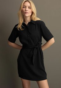 Schwarzes Hemdkleid mit kurzen Ärmeln, einer durchgehenden Knopfleiste, einer Brusttasche und einem passenden Stoffgürtel, der in der Taille gebunden ist. Glatte Textur.