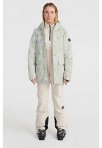 Veste de ski en camouflage vert pâle et blanc, avec une capuche, plusieurs poches et des poignets réglables. Associée à un pantalon de ski beige et des chaussures.