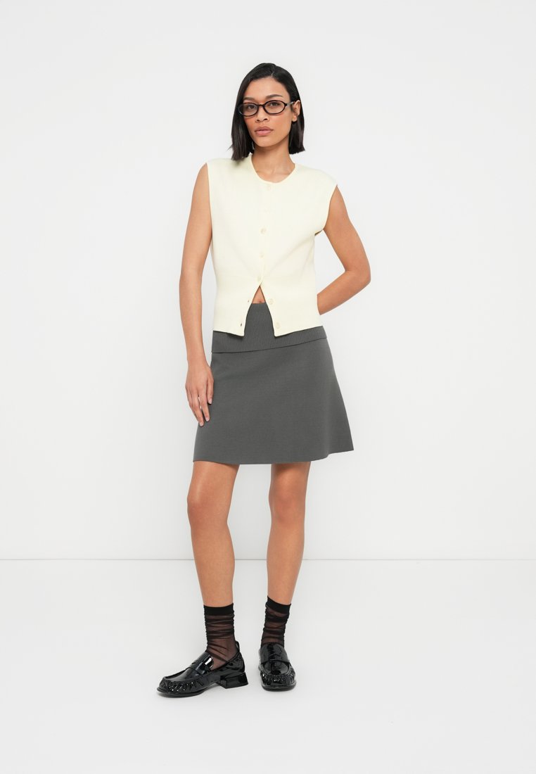 Pull sans manches crème avec boutons, assorti à une jupe trapèze grise. Chaussures noires et chaussettes rayées complètent la tenue. Design minimaliste.