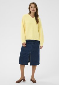 Pull en maille côtelée jaune avec un col en V, associé à une jupe en denim bleu foncé avec des rayures verticales et une fente à l'avant. Chaussures à talons marron.