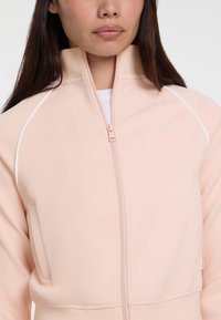 Lichtroze zip-up jas met een gestructureerde afwerking, witte accenten langs de schouders en een logo op het borstzakgedeelte.