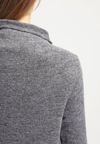 Šedý pletený svetr s jemnou texturou, který má límec a jednoduchý design. Pohled zezadu ukazuje hustou pleteninu a neutrální odstín.