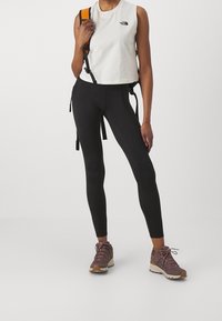 Leggings neri e un top cropped bianco con logo. Il modello indossa scarpe da trekking marroni e porta uno zaino con dettagli arancioni.