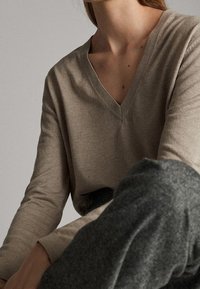 Personne portant un pull beige à col en V et manches longues et un pantalon gris foncé, assise devant un fond neutre uni.
