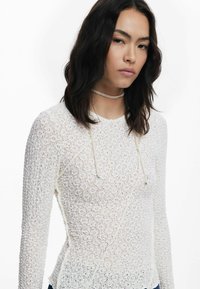 Haut en dentelle blanche à manches longues avec un motif floral texturé, présentant un col rond et des accents à cordon sur le devant.