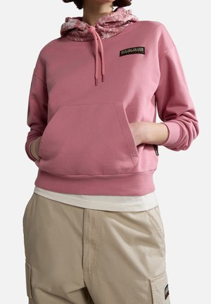 Person trägt rosa Hoodie mit floralem Innenfutter der Kapuze und beige Cargohosen, Hände in der vorderen Tasche.