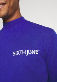 Lila stickad polotröja med en ribbad krage och broderad "SIXTH JUNE" logotyp i vitt på vänster sida. Slät textur.