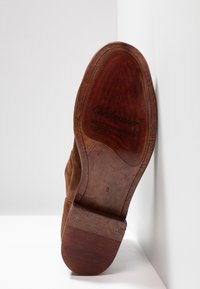 Brązowa zamszowa podeszwa buta z drewnianym obcasem o gładkim wykończeniu, wytłoczonym logo marki oraz szczegółami konstrukcji Goodyear welt.