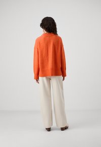 Oransje strikkegenser med avslappet passform, rund halslinning og ribbestrikkede mansjetter. Kombinert med vide beige bukser og brune blokkhæl sko.