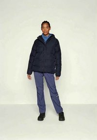 Moteris stovi veidu į priekį, vilkinti juodą pūkinę striukę, mėlyną megztinį su aukštu kaklu, tamsiai mėlynas kelnes ir juodus batus, prieš paprastą šviesią fondo.